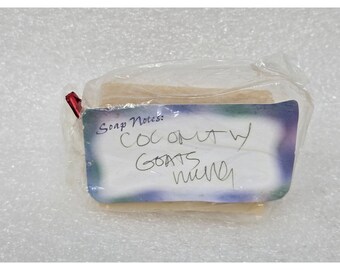May include: A bar of soap wrapped in clear plastic. The soap has a label that reads "Soap Notes: Coconut w/ Goats Milk". The label has a blue and purple border. A red tag is attached to the plastic.