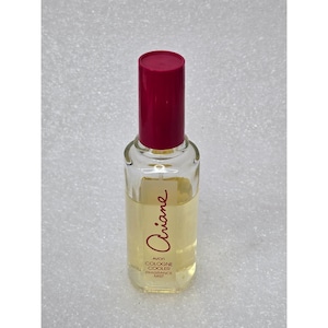 May include: A clear glass perfume bottle with a red cap. The bottle contains a light yellow liquid and has the name "Ariane" written in cursive. The bottle also has the text "Avon Cologne Cooler Fragrance Mist".