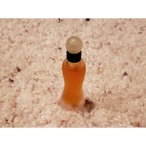 May include: A small, hourglass-shaped glass perfume bottle filled with an amber-coloured liquid. The bottle features a black neck collar and a smooth, round, opaque white cap that sits securely on top of the narrow opening of the glass container.