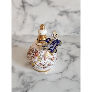 Può includere: Un flacone di profumo decorativo e ornato con un design bianco, oro e floreale. È attaccata un'etichetta blu a forma di farfalla con la parola "PASSION". Il flacone ha un collo dorato e un tappo bianco.