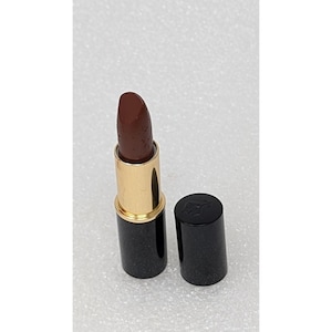 Puede incluir: Un labial marrón oscuro con una base dorada y una tapa negra. El labial está abierto, revelando el color. La tapa se coloca junto al labial.