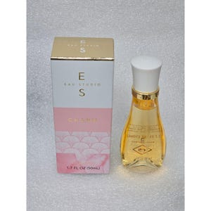 May include: A glass perfume bottle with a white cap and golden liquid, accompanied by its rectangular box. The packaging features the text EAU STUDIO, CHARM, and 1.7 FL OZ (50mL) in gold lettering against white, pink, and patterned sections.