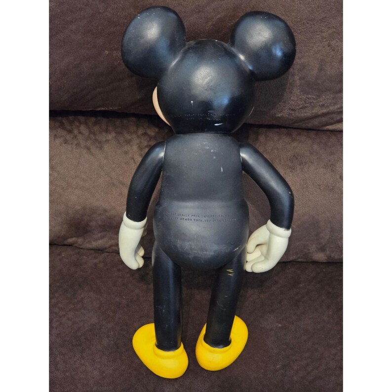 May include: A vintage Mickey Mouse figurine, viewed from the back. The figure is primarily black with white gloves, yellow shoes, and a tan face. The ears are large and round. The figure is standing.