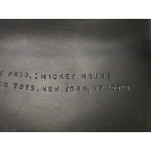 May include: Close-up of a dark gray surface with embossed text. The text reads "PROD.: MICKEY MOUSE" and "GO TOYS, NEW YORK, NY 10019."