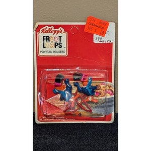 Op de afbeelding: Een verpakking Froot Loops haarbandjes met drie tukan-figuurtjes met rode, blauwe en gele accenten. De verpakking is rood en wit met het Kellogg's-logo en de tekst "Froot Loops Ponytail Holders".
