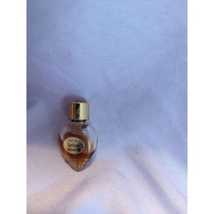 May include: A small, heart-shaped perfume bottle with a gold cap. The clear glass bottle contains a light brown liquid and has a label that reads "AVON Cotillion Perfume Oil."