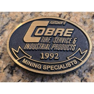 May include: An oval-shaped, two-tone metal plaque with the text "Fletcher's COBRE TIRE, SERVICE & INDUSTRIAL PRODUCTS 1992 MINING SPECIALISTS". The text is in gold against a black background.