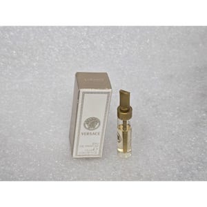 May include: A small Versace Eau de Parfum sample vial with a gold cap and a matching white rectangular box. The box features the Versace logo, the text "VERSACE EAU DE PARFUM", and the volume "1.8 ml e 0.06 US FL. OZ." printed in black.