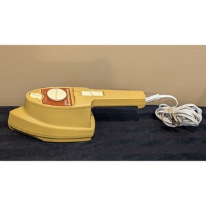 May include: A beige handheld fabric steamer with a white dial and a white cord. The steamer has the text "Sunbeam" printed on the side.