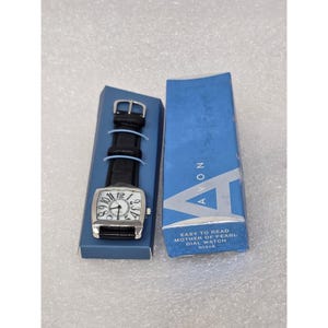 May include: A black leather strap watch with a rectangular silver face and large white numbers. The watch is in a blue box with the Avon logo and text that reads "Easy to Read Mother of Pearl Dial Watch black."