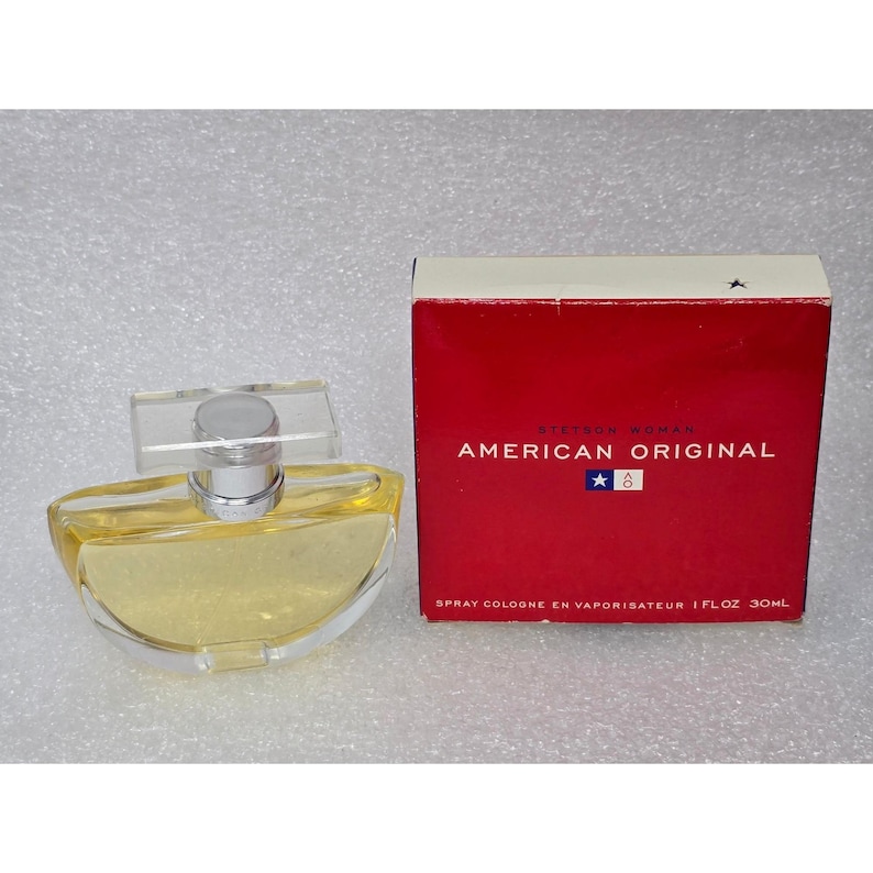 May include: A clear glass perfume bottle with a silver cap and a rectangular clear top, next to a red box. The box has white text that reads "STETSON WOMAN AMERICAN ORIGINAL" and "SPRAY COLOGNE EN VAPORISATEUR 1 FL OZ 30ML".