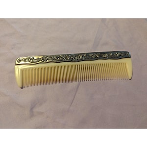 May include: A vintage comb featuring a cream-coloured body and fine teeth. The top edge is decorated with an ornate silver-toned metal design. This antique hair accessory is a classic example of early 20th-century craftsmanship.