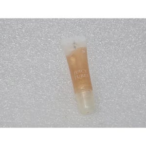 Puede incluir: Un pequeño tubo de plástico transparente de brillo labial Lancôme Juicy Tubes. El producto contiene una fórmula líquida dorada y brillante, con el nombre de la marca y el texto JUICY TUBES impresos en blanco en la parte frontal del tubo.