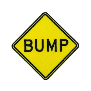 Könnte beinhalten: Gelbes rautenförmiges Abzeichen mit schwarzem Rand und dem Wort „BUMP“ in fetten schwarzen Buchstaben. Das Abzeichen ist ein Verkehrsschild-Design, das üblicherweise verwendet wird, um eine Unebenheit auf der Straße anzuzeigen.