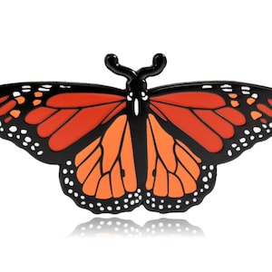 May include: A monarch butterfly-shaped decorative item. The butterfly has orange and black wings with white spots along the edges. The body is black with two antennae. The item is likely a pin or a magnet.