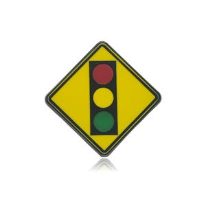 Puede incluir: Un pin en forma de rombo con fondo amarillo y borde negro. Centrado en el pin hay un panel rectangular negro vertical que contiene tres luces circulares en una configuración de semáforo: rojo arriba, amarillo en el medio y verde abajo.