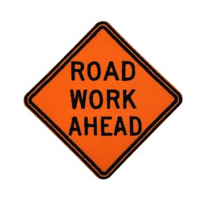 Könnte beinhalten: Ein orangefarbenes, rautenförmiges Verkehrsschild mit schwarzem Text „ROAD WORK AHEAD“. Das Schild hat einen schwarzen Rand und warnt Verkehrsteilnehmer vor Straßenbauarbeiten.