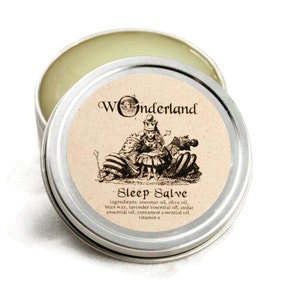 May include: A silver tin with a label that reads "Wonderland Sleep Salve" and lists the ingredients: coconut oil, olive oil, beeswax, lavender essential oil, cedar essential oil, cinnamon essential oil, and vitamin E. The label features a black and white illustration of three figures from Alice in Wonderland.