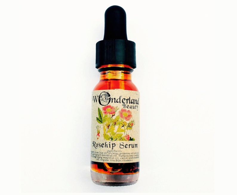 May include: A small amber glass bottle with a black dropper top. The bottle is labeled "Wonderland Beauty Rosehip Serum" and contains a light orange liquid. The label features a botanical illustration of a rosehip plant with pink flowers.