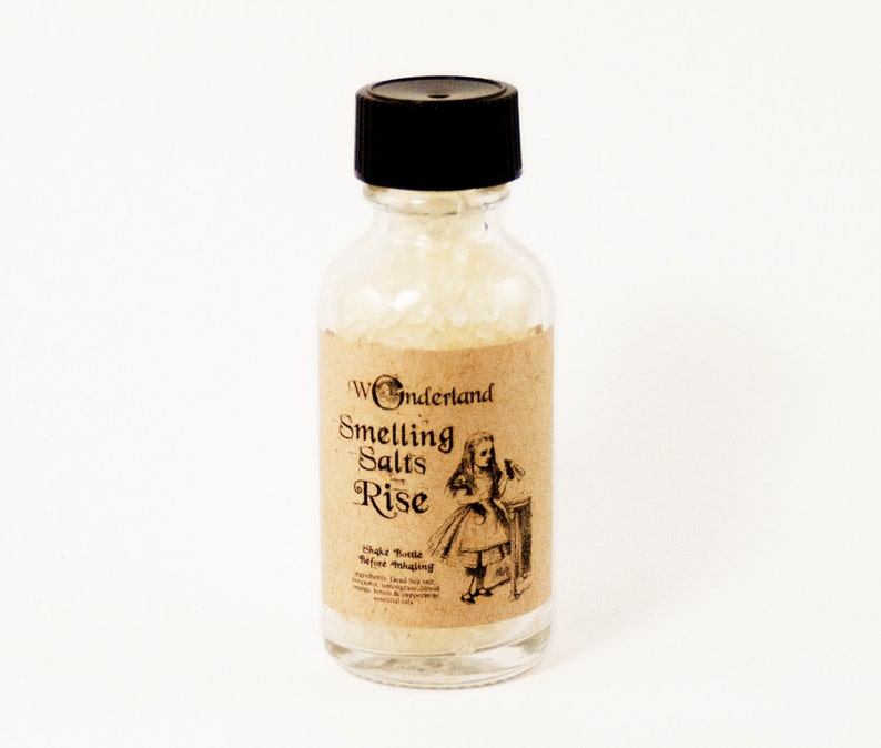 May include: A small glass bottle with a black lid filled with white smelling salts. The label is brown with white text that reads "Wonderland Smelling Salts Rise" and features a black and white illustration of Alice in Wonderland.