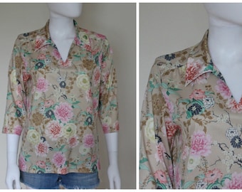 Puede incluir: Blusa color beige con estampado floral en rosas, blancos y verdes. Tiene cuello en V y mangas tres cuartos. La tela parece ligera. La blusa se muestra en un maniquí.
