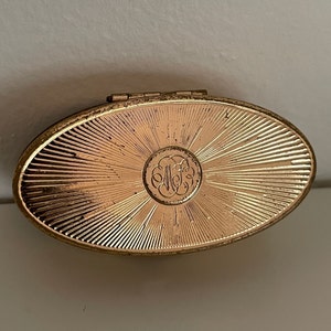 May include: Oval-shaped, gold-toned compact with a sunburst design radiating from a central medallion. The medallion features an ornate monogram. The compact has a hinged lid and a slightly worn appearance.