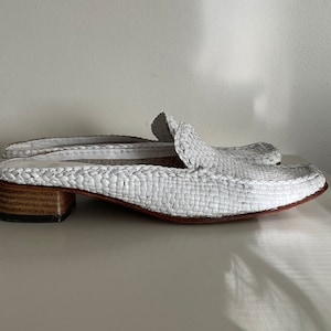 May include: A pair of white woven leather mules with a square toe and low, stacked heel. The shoes have a slip-on design and a textured, basket-weave pattern. The heels are approximately 3.8 cm high.