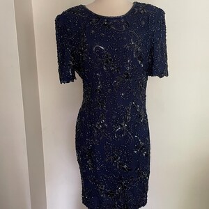 May include: A navy blue, short-sleeved dress with intricate sequin and bead embellishments. The dress features a round neckline and a fitted silhouette, showcasing a detailed floral pattern. The dress is displayed on a mannequin.