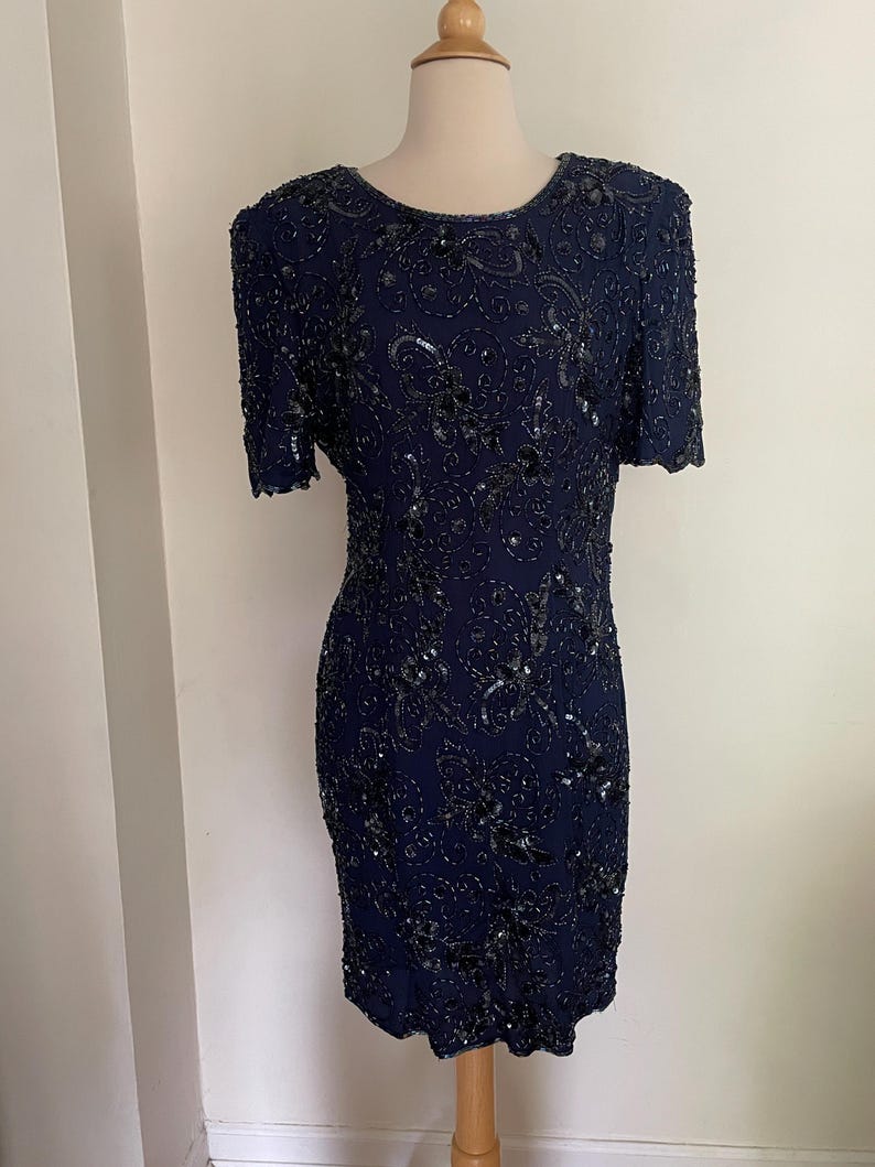 May include: A navy blue, short-sleeved dress with intricate sequin and bead embellishments. The dress features a round neckline and a fitted silhouette, showcasing a detailed floral pattern. The dress is displayed on a mannequin.