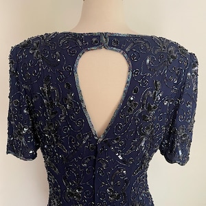 May include: Navy blue dress with short sleeves, adorned with intricate black sequin and bead embellishments. The dress features a keyhole back detail and a subtle floral pattern, creating a sophisticated and elegant look. The dress is displayed on a mannequin.