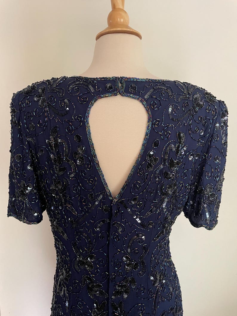 May include: Navy blue dress with short sleeves, adorned with intricate black sequin and bead embellishments. The dress features a keyhole back detail and a subtle floral pattern, creating a sophisticated and elegant look. The dress is displayed on a mannequin.