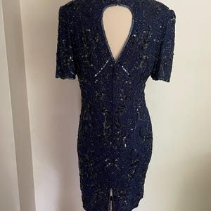 May include: A navy blue, short-sleeved dress with intricate sequin and bead embellishments. The dress features a keyhole back and a slit at the hem. The dress is displayed on a mannequin.