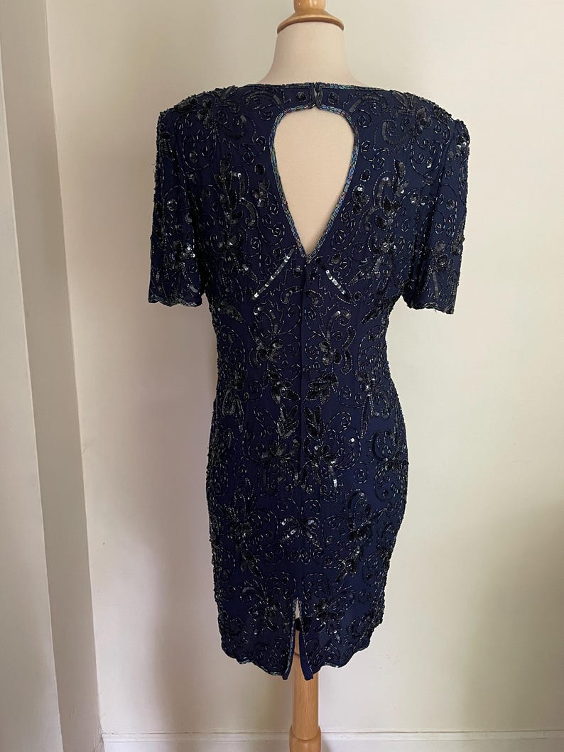 May include: A navy blue, short-sleeved dress with intricate sequin and bead embellishments. The dress features a keyhole back and a slit at the hem. The dress is displayed on a mannequin.