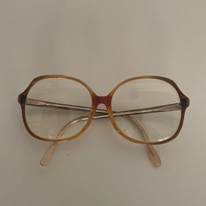 May include: Vintage eyeglasses with a brown and tan frame. The glasses have a rounded, oversized shape with clear lenses and a metal bridge. The temples are a light tan color. The glasses are set against a plain white background.
