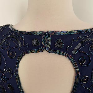 May include: Navy blue beaded dress with a keyhole back. The dress features intricate beadwork and sequin embellishments around the neckline and keyhole. The beads have iridescent colors. The dress is displayed on a mannequin.