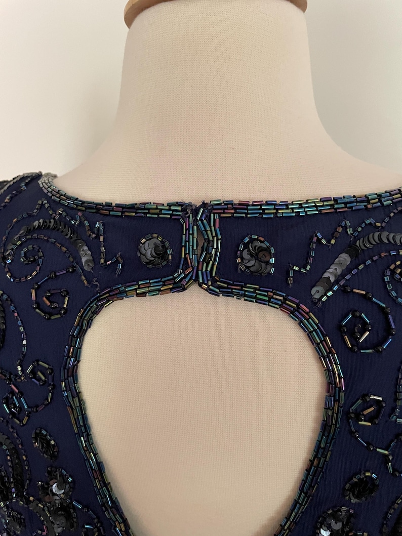 May include: Navy blue beaded dress with a keyhole back. The dress features intricate beadwork and sequin embellishments around the neckline and keyhole. The beads have iridescent colors. The dress is displayed on a mannequin.