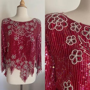 Puede incluir: Top rojo con lentejuelas con motivos florales y de hojas. La parte superior de manga larga presenta un dobladillo festoneado y está adornada con detalles de cuentas blancas. El diseño incluye un cuello redondo y se muestra en un maniquí.