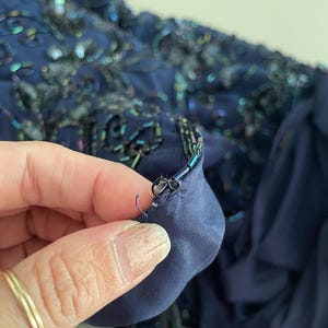 May include: Close-up of a navy blue fabric with intricate bead and sequin embellishments. The fabric has a layered texture, with a darker blue lining visible. The details include iridescent beads and sequins.