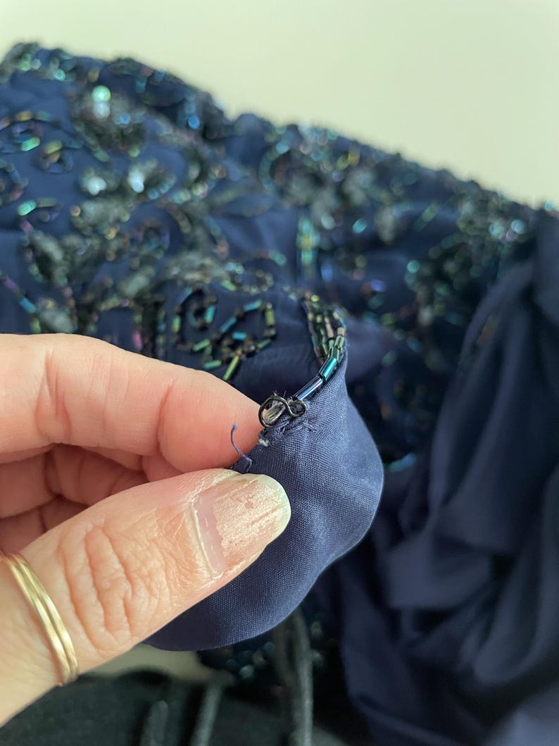 May include: Close-up of a navy blue fabric with intricate bead and sequin embellishments. The fabric has a layered texture, with a darker blue lining visible. The details include iridescent beads and sequins.