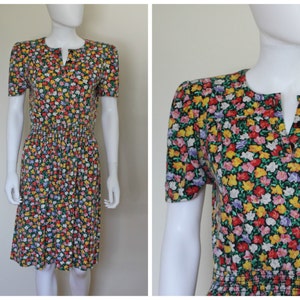 May include: A vintage floral print dress with a dark green background. The dress features a V-neckline, short sleeves, and a gathered waist. The colorful floral pattern includes red, yellow, and pink flowers. The dress is knee-length.