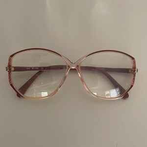 May include: A pair of vintage eyeglasses with a light pink frame and dark brown temples. The lenses are clear, and the frame has a butterfly shape. The text "SPX M 1793 720" is visible on the frame.