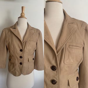 May include: A tan leather jacket with three-quarter sleeves, a notched collar, and two front pockets. The jacket has dark brown buttons and decorative stitching details. The jacket is displayed on a mannequin.
