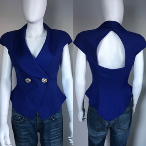 May include: A royal blue sleeveless blazer with a double-breasted design. The blazer features two decorative rhinestone buttons and a unique open back. The garment is displayed on a mannequin.
