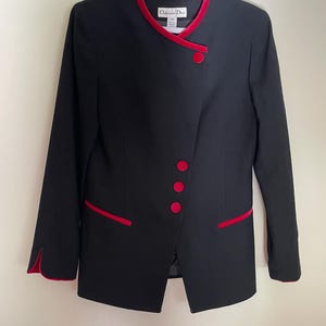 Puede incluir: Blazer negro con ribete y botones de terciopelo rojo. El blazer tiene un diseño asimétrico único con cuello con muescas y mangas largas. La etiqueta Christian Dior es visible.