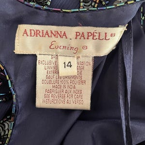 May include: Close-up of a navy blue evening gown tag. The tag reads "ADRIANNA PAPELL Evening" and includes size 14. The gown is embellished with sequins and beads. The tag also includes care instructions and material information.