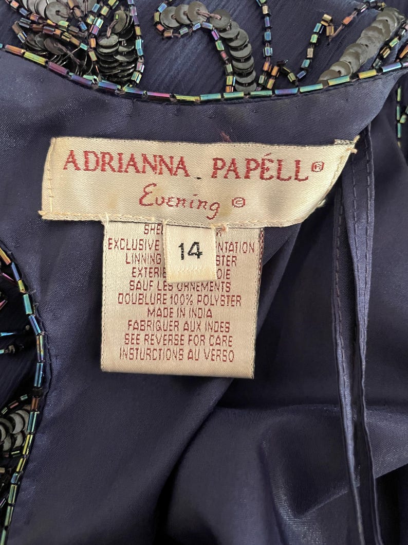May include: Close-up of a navy blue evening gown tag. The tag reads "ADRIANNA PAPELL Evening" and includes size 14. The gown is embellished with sequins and beads. The tag also includes care instructions and material information.