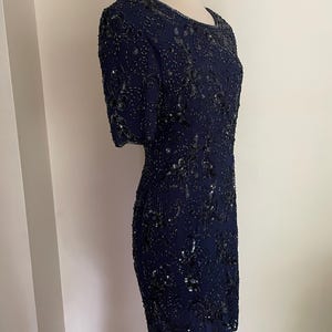 May include: A navy blue, knee-length dress with short sleeves. The dress is covered in intricate black beading and sequins, creating a shimmering floral pattern. The dress is displayed on a mannequin.