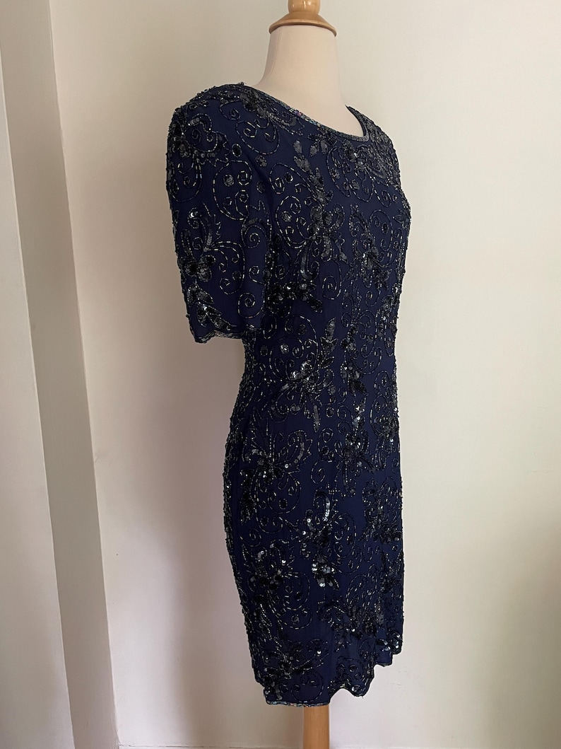 May include: A navy blue, knee-length dress with short sleeves. The dress is covered in intricate black beading and sequins, creating a shimmering floral pattern. The dress is displayed on a mannequin.