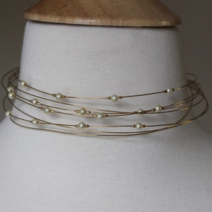 Puede incluir: Un delicado collar gargantilla con múltiples hilos de alambre fino de color dorado. Pequeñas perlas redondas de color blanquecino están espaciadas uniformemente a lo largo de los hilos. El collar se exhibe en un busto de maniquí blanco.