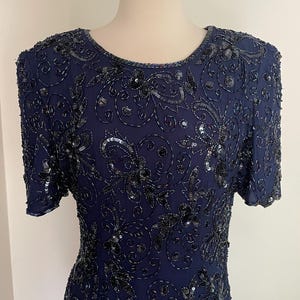 May include: Navy blue dress with short sleeves, embellished with intricate black sequins and beads. The dress features a round neckline and a detailed floral pattern across the bodice and sleeves. The dress is displayed on a mannequin.
